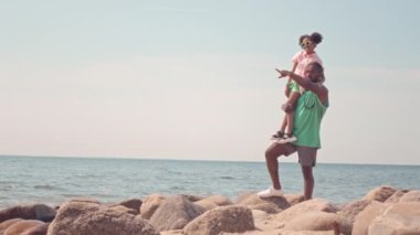 Pembe günbatımında kumsalda taşların üzerinde duran küçük kızı olan uzun boylu, yavaş siyah bir adamın portresi.