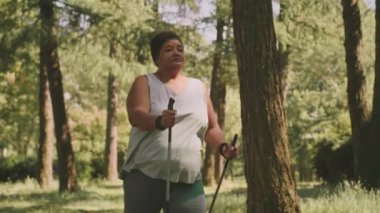 Orta yavaş kıvrımlı olgun siyah kadın yürüyüş direkleri ile yaz günü parkta yürüyor.