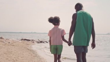 Orta geri görüşlü Afrikalı Amerikalı bir adam ve küçük kızı gün batımında kumsalda el ele yürürken görülüyor.