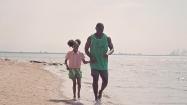 Gün batımında kumlu deniz kıyısında koşan küçük kızı olan yakışıklı siyahi bir adam.