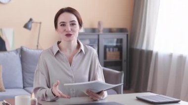 Elinde bir yığın iş belgesi olan beyaz iş kadınının belini kaldırın. Kapalı bir masada oturuyor ve kameraya konuşuyor.