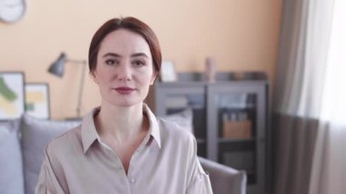 Beyaz saçlı genç bir kadının göğüs portresi. Ofiste kameraya gülümsüyor. Şeftali rengi duvarlar ve gri mobilyalarla.