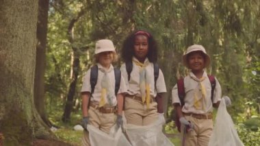 Ortaçağ çocuklarından oluşan çok ırklı izci grubunun orta boy portresi güneş ışığında yeşil yaz ormanlarında çöp torbalarıyla durup çevreye yardım ediyor.