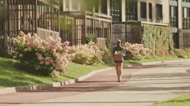 Kutuda yiyecek taşıyan genç siyahi bir adamın güneşli bir günde modern apartman bölgesinde yürüyüp müşterilerine yiyecek götürdüğü uzun bir çekim.