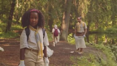 Ormanda kameraya poz veren küçük Afro-Amerikalı kızın orta boy portresi. Bu arada izci lideri kafasında arka planda çöp topluyor.