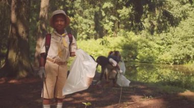 Elinde çöp torbasıyla küçük Afro-Amerikan izci çocuğun orta boy portresi sürüsüyle birlikte ormanı temizlemeye yardım ediyor.