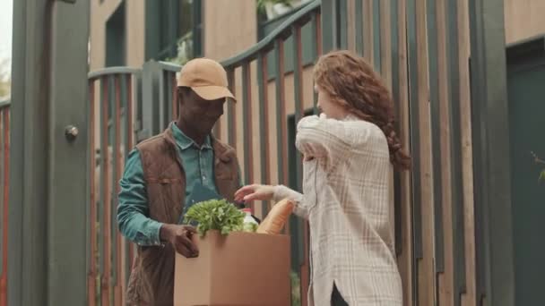 若い感謝する女性に食料品を届ける若い黒人男性宅配便のミディアムショット 彼女の現代的な建物のアパートの屋外の門に立って — ストック動画
