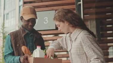 Kafkasyalı genç bir kadının market ürünleriyle karton kutunun içine bakarken orta boy fotoğrafı. Genç Afrikalı Amerikalı erkek kuryenin siparişlerini kontrol ederken.
