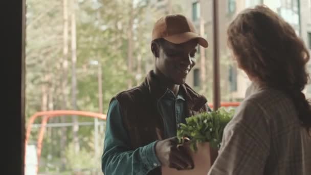 中镜头 年轻的非洲裔美国男性信使在家里将装有食品杂货的盒子交给用智能手机支付订单的年轻白人妇女 — 图库视频影像