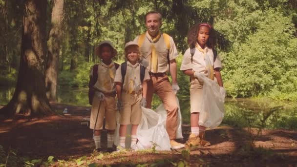 ゴミからそれを収集した後 日光の緑の森に立っているカメラのためにポーズするゴミ袋を持つ多民族の子供の部隊を持つ大人の白人男性のスカウトリーダーの全長のスローモの肖像画 — ストック動画