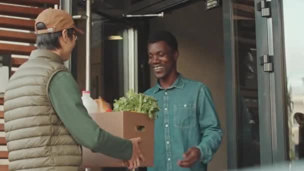 エントランスドアでアジアの急使ミーティングから食料品の配達を受け取る陽気な若い黒人のミディアムショット — ストック動画