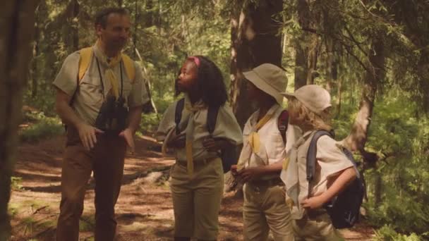 大人の白人男性スカウトリーダーとスカウトユニフォームのマルチエスニック子供のグループを撮影し 晴れた日に一緒に森林エリアを探索 — ストック動画