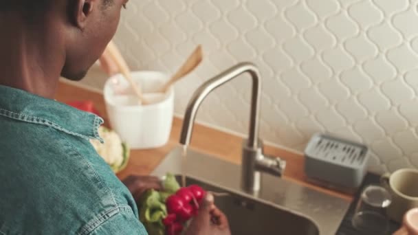 穿着斜纹棉布衬衫的年轻非洲裔美国人肩膀上的照片 他在家里做饭的时候用水槽洗新鲜蔬菜 — 图库视频影像