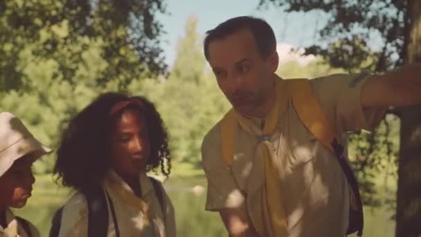 ツリーのトランクにリーニングし スカウトユニフォームでアフリカ系アメリカ人の子供のグループに森の植物について話す成人男性の白人スクーターの中型ショット — ストック動画