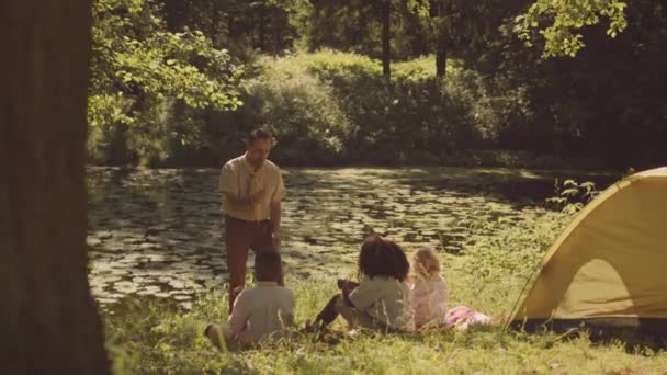 大人の白人男性のスカウトリーダーは 日光で照らされた緑の森のグレードに座っている子供のキャンプスキルの多様なグループを教えています — ストック動画