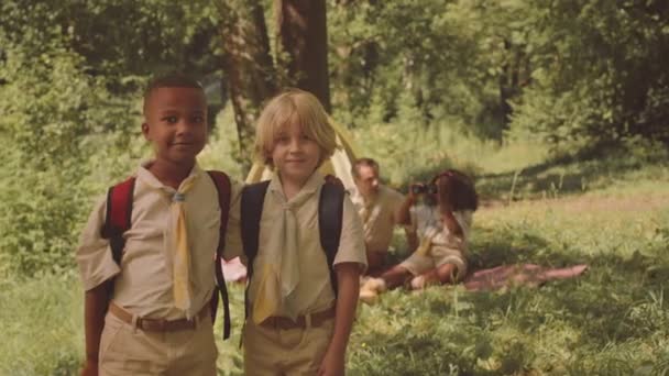 2つの多様な小学生のボーイスカウトの中央スローモの肖像画は 日光の森のグレードのテントに対して立っているカメラのためにポーズし 夏に軍隊と彼らのスカウトリーダーと一緒にキャンプ — ストック動画