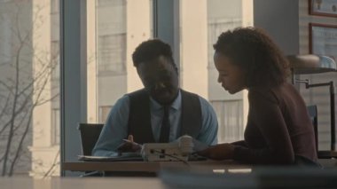 Genç ve başarılı Afro-Amerikan iş ortakları modern ofiste büyük pencereli masa başında konuşuyor.