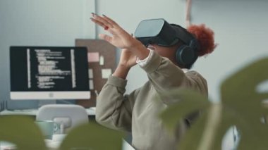 Genç Afrikalı Amerikalı yazılım mühendisi ellerini havada hareket ettirirken kapalı alanda gözlük takarak sanal gerçekliği deneyimliyor.