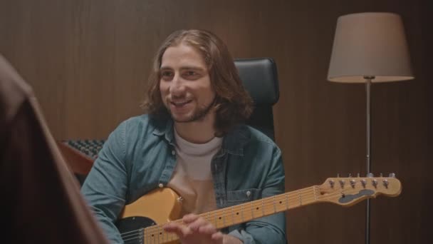 stock video Young long haired musician with electric guitar talking to his music producer while recording album at professional record studio