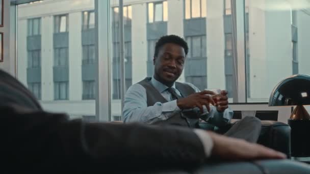 パノラマウィンドウ付きラグジュアリーオフィスでブラックレザーコーチに座っている彼のビジネスパートナーと手でウイスキーのグラスと自信を持ったアフリカ系アメリカ人のビジネスマンのミディアムショット — ストック動画