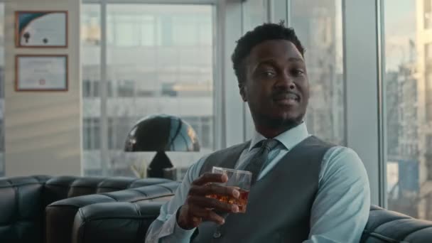 若い成功したアフリカ系アメリカ人のビジネスマンのミディアム肖像画 彼の豪華なオフィスで黒い革のソファに座っているウイスキーのガラスとカメラをポーズ — ストック動画