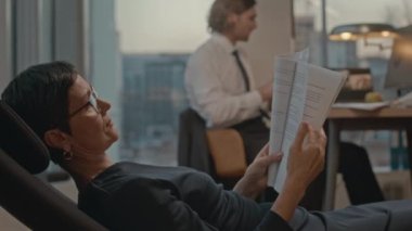 Olgun Kafkasyalı iş kadını, panoramik pencereler okuma sözleşmesi ile ofisinde sandalyeye yaslanıyor.