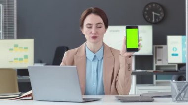 Beyaz tenli genç iş kadınının belinde boş bir akıllı telefon ekranı beliyle ofiste oturuyor ve kameraya konuşuyor.