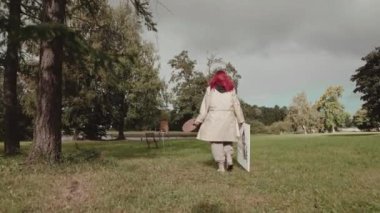Beyaz tuval ve boyalarla açık havada resim yapmak için yaz parkına giden genç kızıl saçlı kadın ressamın arka görüntüsü.