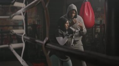 Güçlü, genç Müslüman bir kadının boks eldivenleriyle kum torbasına vururken kapalı alanda antrenman yaparken orta boy fotoğrafı.