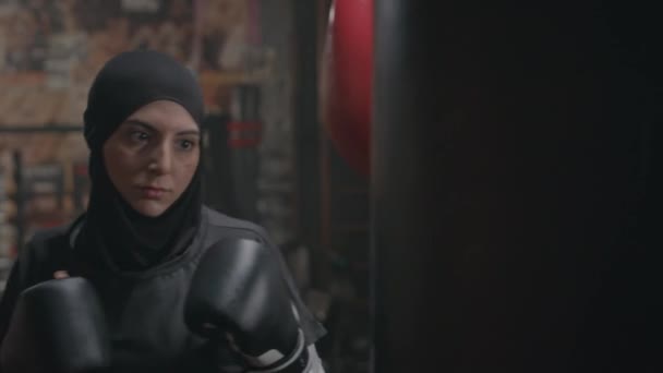 穿着黑色头巾和拳击手套的年轻穆斯林女子在室内拳击训练中击球袋的决心 — 图库视频影像