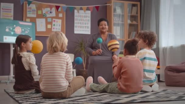 在明亮的教室里 一群有着太阳系Diy行星的孩子手牵着手围坐在地板上 与黑人女教师一起上互动科学课 — 图库视频影像