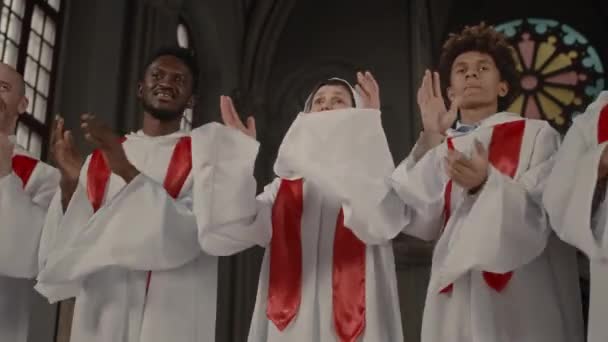 長い白いガウンを身に着け 古いカトリック教会で一緒に立って一緒に歌う喜びに満ちた多民族教会合唱団の低角度媒体ショット — ストック動画
