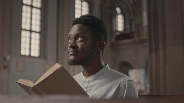 カトリック教会のベンチに座っている間 聖書を読んで神に頭を上げる若いアフリカ系アメリカ人のチェストアップ — ストック動画