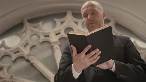 教堂礼拜时 中青年秃头天主教牧师站在玫瑰窗下读圣经的低角度 — 图库视频影像