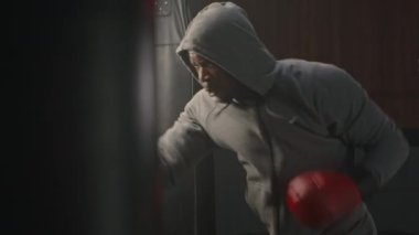 Gri spor giyim ve kırmızı eldivenli agresif siyah boksör içeride antrenman yaparken kum torbasına vuruyor.