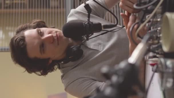 スタジオで空中でライブしている間にマイクで話す若いコーカサスラジオホストの垂直ウエストアップショット — ストック動画