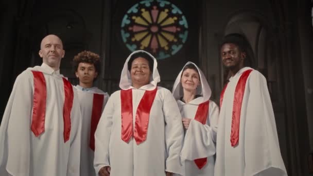 白いガウンの多様な教会合唱団メンバーの中央の肖像画は 背景にステンドグラスローズウィンドウで古いカトリック教会に立っているカメラのために一緒にポーズします — ストック動画