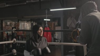 Siyah başörtüsü ve spor giyimli genç Müslüman kadın ringde boks antrenmanı yaparken siyah erkek koçuyla antrenman yapıyor.