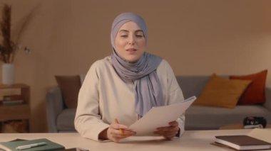Elinde bir yığın belge tutan ve oturma odasındaki masada oturan kameraya konuşan tesettürlü genç Müslüman kadının belini kaldır.