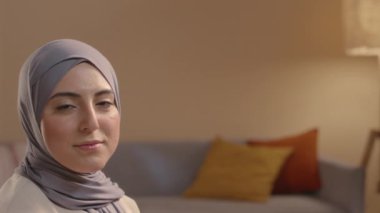 Sıcak oturma odasında kameraya poz veren tesettürlü gülümseyen Müslüman kadın portresi.