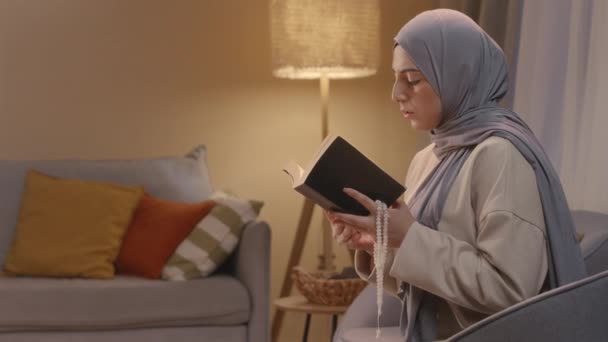 头戴头巾的年轻穆斯林妇女坐在舒适的客厅里的椅子上 手里拿着米赫巴哈 正在阅读 古兰经 — 图库视频影像