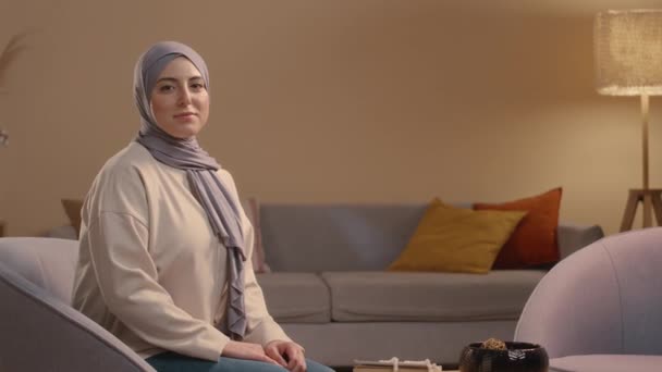 坐在舒适的客厅里 坐在椅子上 头戴头巾 对着相机微笑的年轻穆斯林妇女的中等画像 旁边放着可兰经和米沙咖啡桌 — 图库视频影像