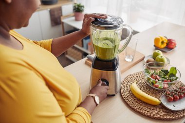 Ev mutfağında sağlıklı meyve püresi yaparken aşırı kilolu siyahi bir kadının blender kullanmasının yüksek açılı yakın çekimi.