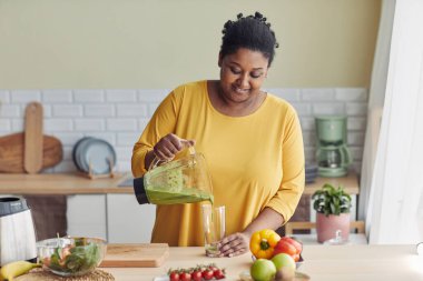 Şişman siyahi kadın portresi ev mutfağında sağlıklı meyve suyu içiyor, boşluğu kopyalıyor.
