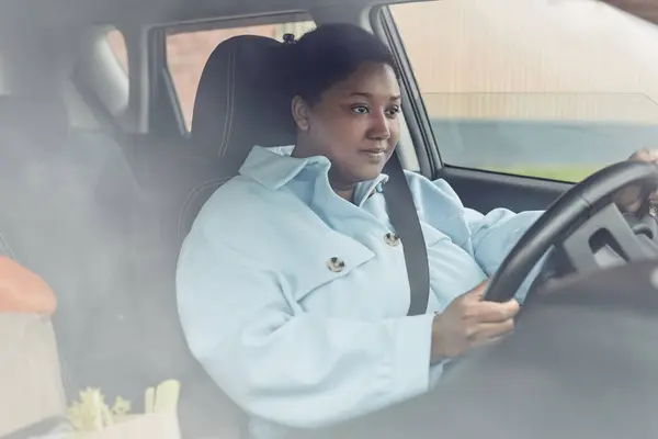Behind glass view of young black woman driving car with seatbelt on, copy space