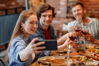 Sıcak bir ortamda yemek masasında arkadaşlarıyla selfie çeken gülümseyen genç bir kadının portresi.