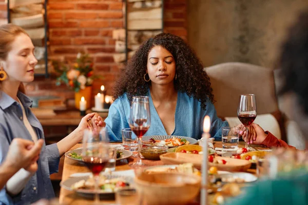 Portrait of young black woman holding hands with friends and saying grace at dinner party in cozy setting, eyes closed