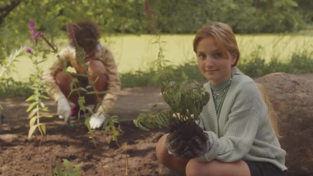 カメラで笑顔のコーカサス女の子の肖像画は 環境を助けるためのエコプロジェクトのために土壌に植える準備を手で緑の植物と一緒に公共の公園でスカウトしながら — ストック動画