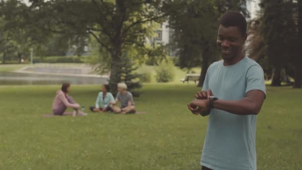 若い黒人男性アスリートのミディアムショット 笑顔ながら公共の公園で芝生に立っているスマートウォッチで屋外のトレーニング — ストック動画