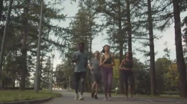 Yeşil parkta koşan dört genç koşucunun düşük açılı tam açılı görüntüsü kameraya doğru ilerliyor.